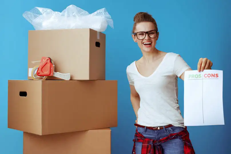 A smiling woman with moving boxes holds a "pros-cons" list, weighing the benefits of commercial cleaning services.