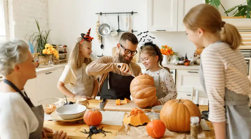 A family carves a pumpkin in a brightly lit kitchen, which will require post-Halloween cleanup tips.