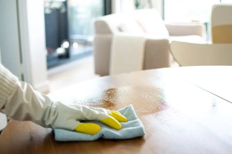 A hand in yellow gloves wipes a wooden table, performing a task on an Airbnb cleaning checklist.