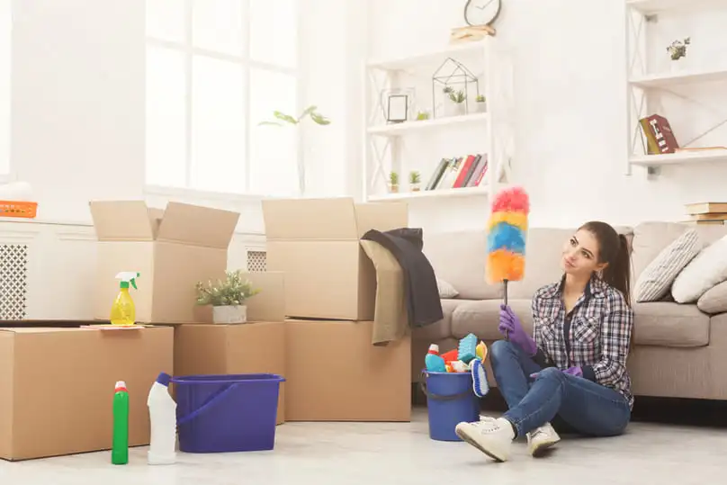 A woman with cleaning supplies and moving boxes, preparing to use a new home cleaning checklist.