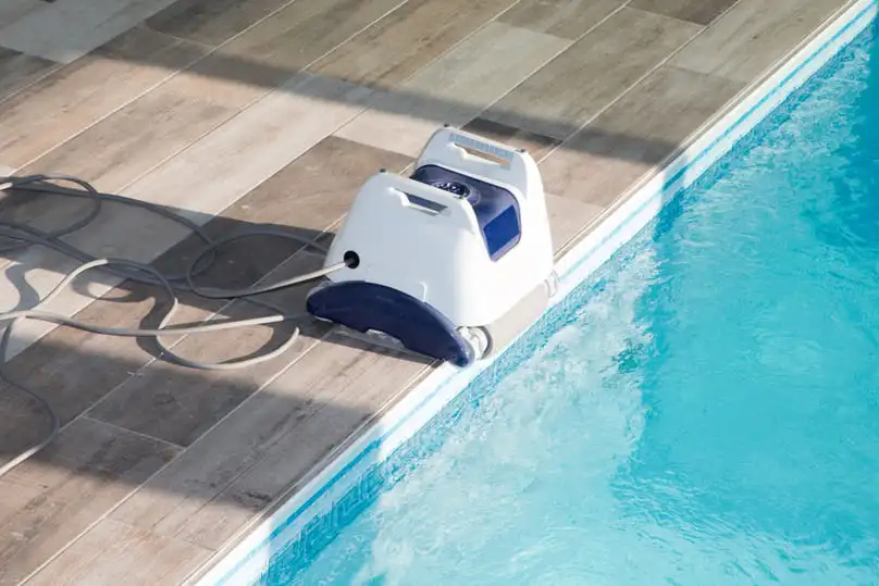 A white and blue robotic pool cleaner sits on the edge of a tiled pool, an example of pool vacuum robots.