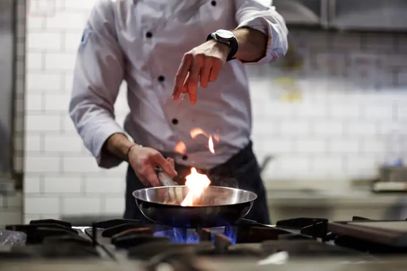 A chef flambés a dish in a hot pan over a gas stove, leading to the need for how to clean a burnt pot.