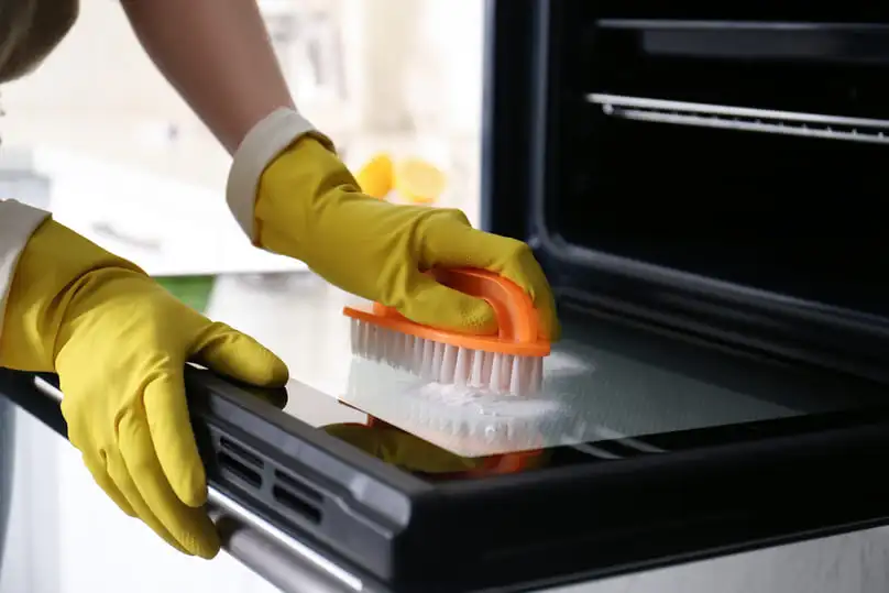 A person in yellow gloves scrubs an open oven door with a brush, showing how to clean oven glass.