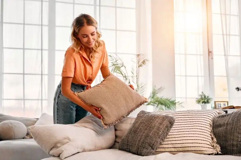 A woman happily arranges throw pillows on a sofa in a sunny room, suggesting how to wash pillows.