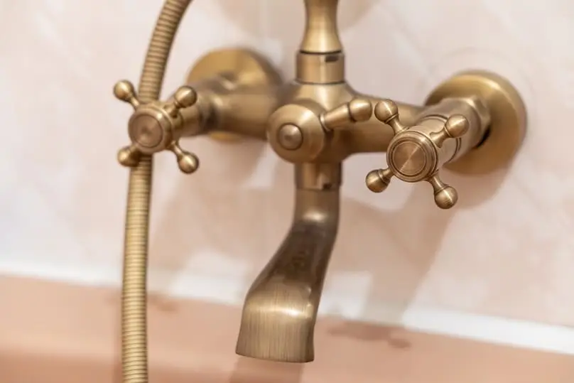 Close-up of a vintage-style brass bathtub faucet with two cross handles and a handheld shower hose, mounted on a light-colored tiled wall.