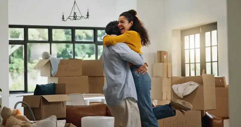 A couple hugs among cardboard boxes in a new home after completing their move out cleaning checklist.
