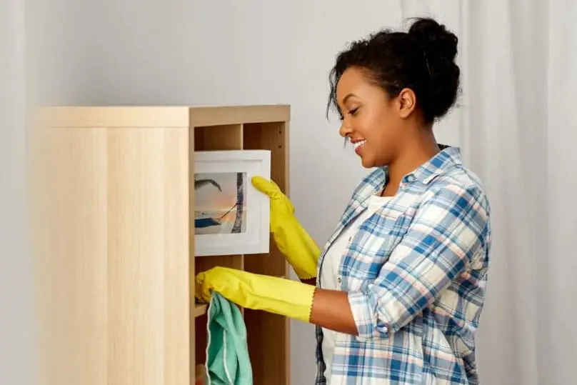 A woman wipes down a wooden bookcase while claning shelves with a green microfiber cloth.