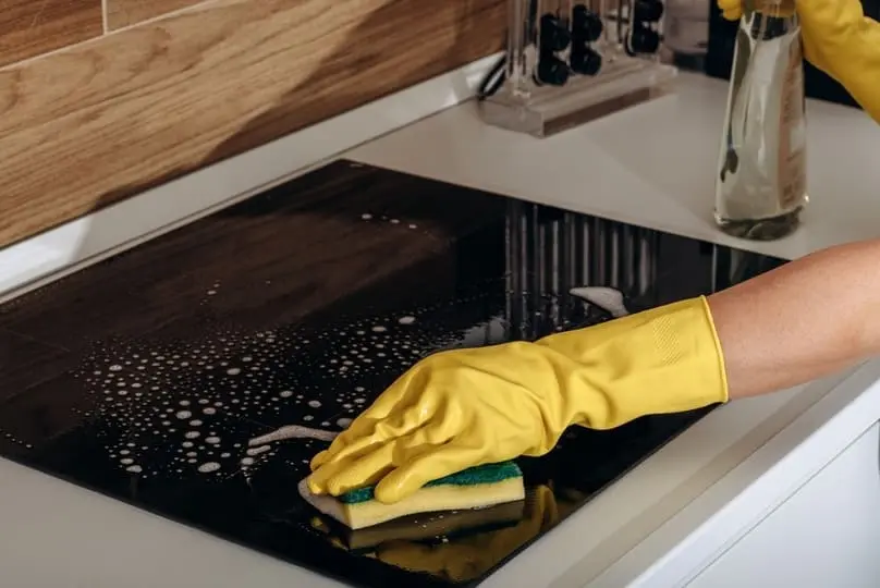 Yellow gloved hands using a sponge show the best way to clean glass cooktop surfaces.