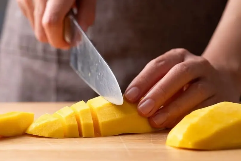 Person slicing a mango on a wooden cutting board before learning how to disinfect cutting boards.