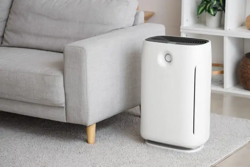 A white air purifier standing by a gray couch shows the best air purifier for allergies.
