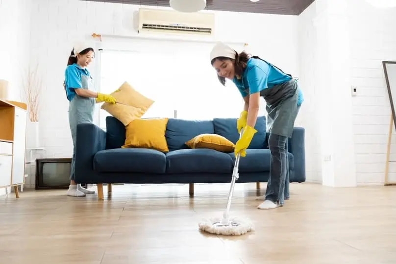 Two professionals mopping and tidying a living room show reasons to hire a house cleaner.